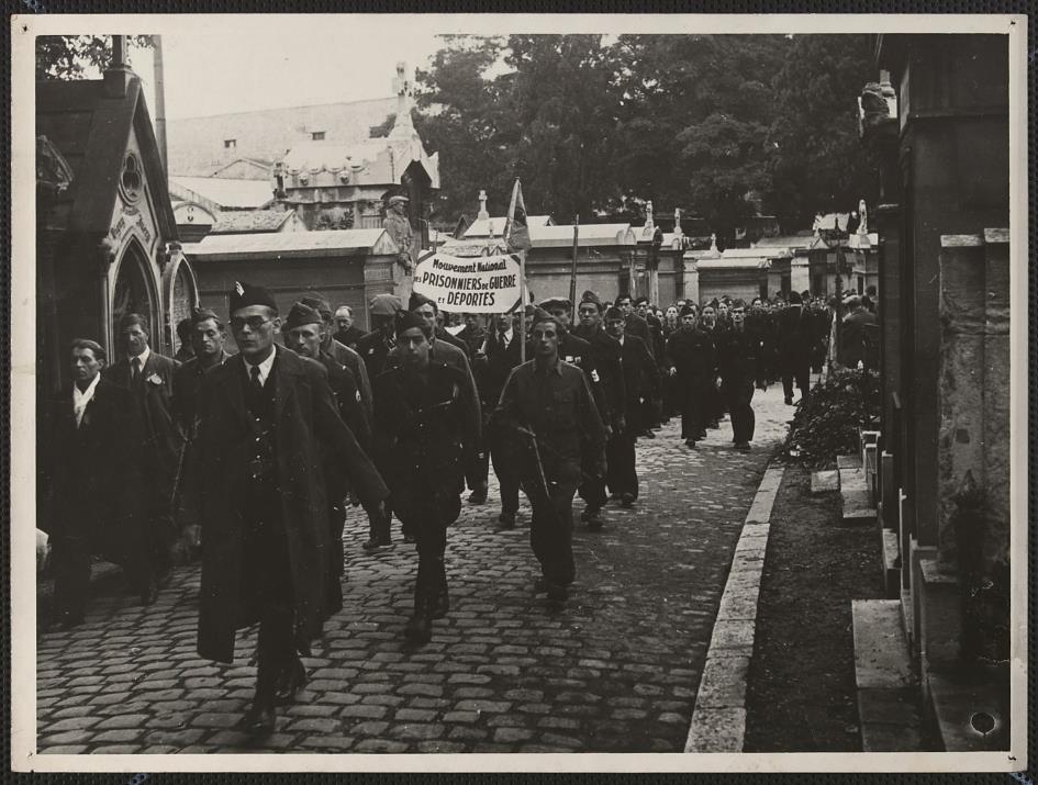 Montée au Mur des Fédérés , défilé de militaires dans les allées du cimetière parisien du Père-Lachaise, certains en armes, avec la banderole « Mouvement national des prisonniers de guerre et déportés ». Paris (75), cimetière du Père-Lachaise, 20e arrondissement, 1944 (Source : Mémoires d'Humanité / Archives départementales de la Seine-Saint-Denis) 