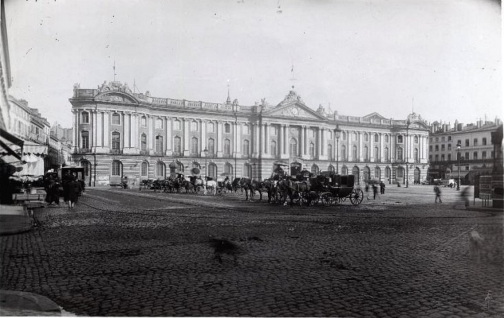 Toulouse (Haute-Garonne), hôtel de ville appelé Capitole. Référence : Place du Capitole. Fin 19e siècle. Vue d'ensemble de la place prise depuis l'angle nord-ouest. Au milieu plusieurs voitures à cheval, au fonds la façade de l'hôtel de ville.  Archives municipales de Toulouse (reproduction), Domaine public.